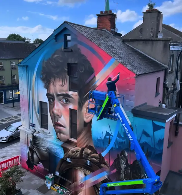 Mister Copy painting Cúchulainn from a blue elevated work platform