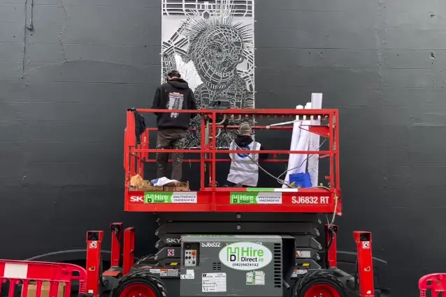 Two artists applying an intricate stencil from a red scissor lift